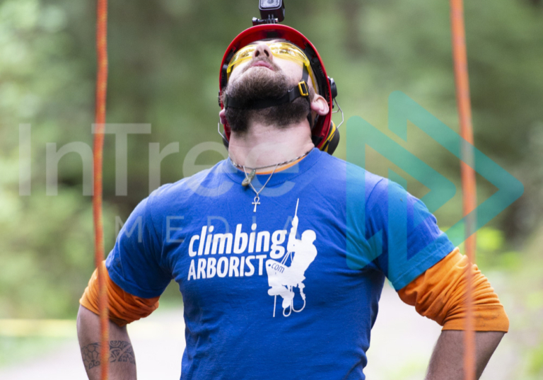 Ground person with GoPro looking up into canopy with two legs of Man wearing helmet, with beard in blue top with hands on hips looking into the forest canopy